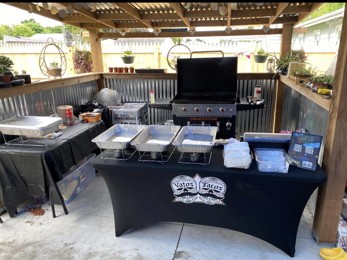 Vatos Tacos branded catering table with chafing dishes set up under a backyard pergola